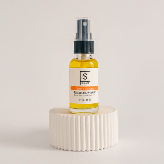 A small white spray bottle labeled SoulDeo Shower Mist with orange and eucalyptus, positioned on a textured white surface against a neutral background.