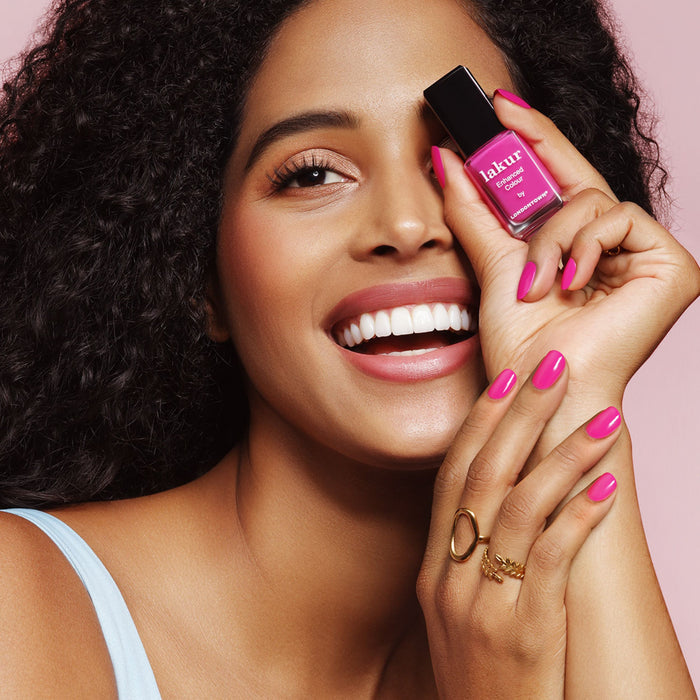 A curly-haired person smiles while holding a bottle of LONDONTOWN's Summer Fling nail polish in a shocking pink shade near their face. The vegan, cruelty-free nails complement their light top perfectly.