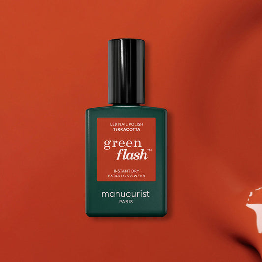 A bottle of Manucurist’s Terracotta gel polish is shown against a red earth–inspired background. The rectangular bottle has a dark green body with a sleek black cap, highlighting the rich Terracotta shade.