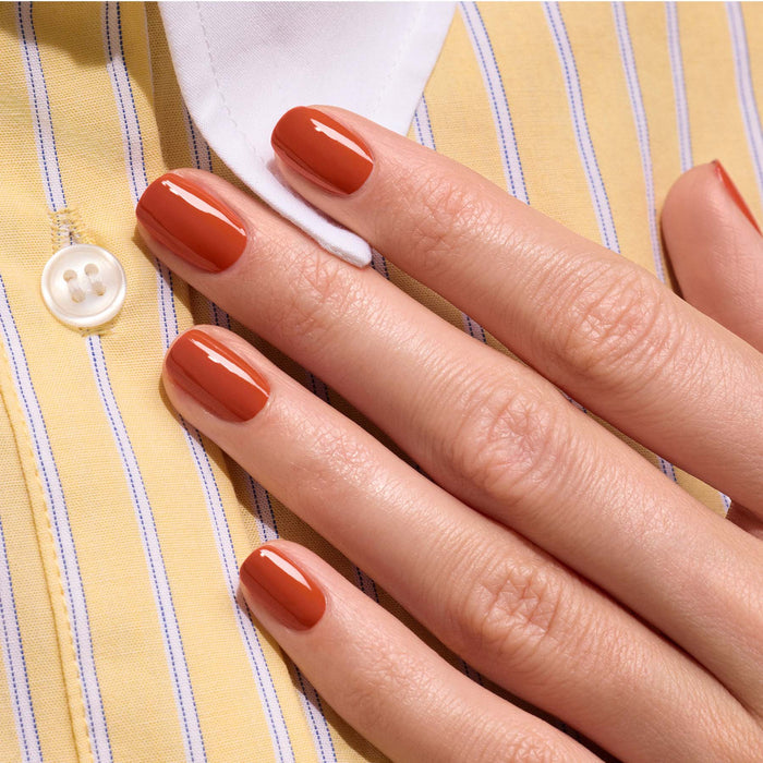 A hand with neatly manicured nails painted in Manucurist’s Terracotta, a glossy scorched red earth shade from the Green Flash gel polish range, rests on a yellow and white striped shirt with a white button.