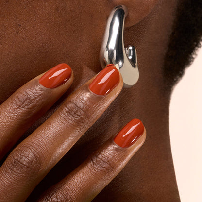 A hand with glossy Terracotta nail polish by Manucurist touches an ear adorned with a chunky abstract silver earring. The neutral background highlights the vibrant polish and striking jewelry.