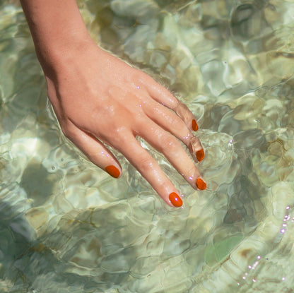 A hand with Terracotta nails, painted using Manucurist's Green Flash gel polish, is gently submerged in clear, sunlit water, creating small ripples and reflections on the surface.
