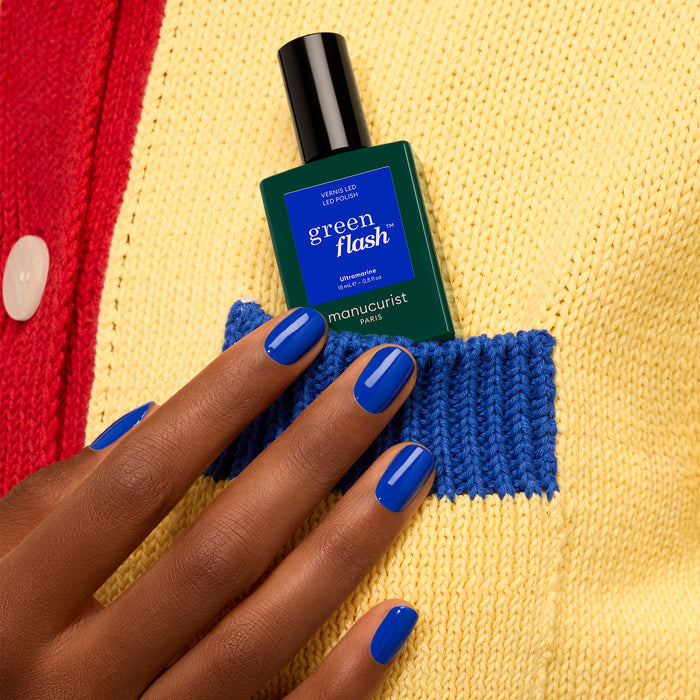 A hand with glossy blue Ultramarine gel polish by Manucurist holds a green Green Flash bottle, tucked into the blue pocket of a yellow and red knit sweater.