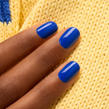 A hand with medium brown skin and short, neatly manicured nails painted in Manucurist's Ultramarine electric blue rests on yellow knit fabric.
