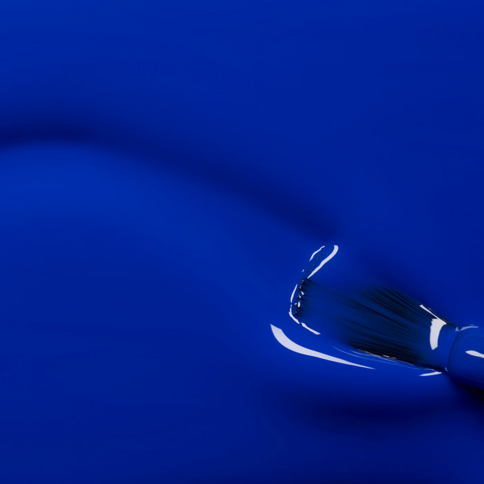 A close-up of a paintbrush applying Manucurist's Ultramarine gel polish, showing glossy reflections on the wet surface and visible bristles for an even, vibrant finish.