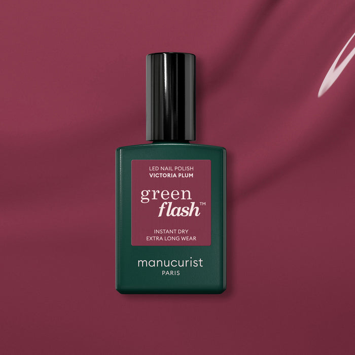 A bottle of Manucurist Victoria Plum gel polish sits on a matching plum background. The sleek dark green bottle has a maroon label and black cap, perfectly highlighting this rich plum shade.