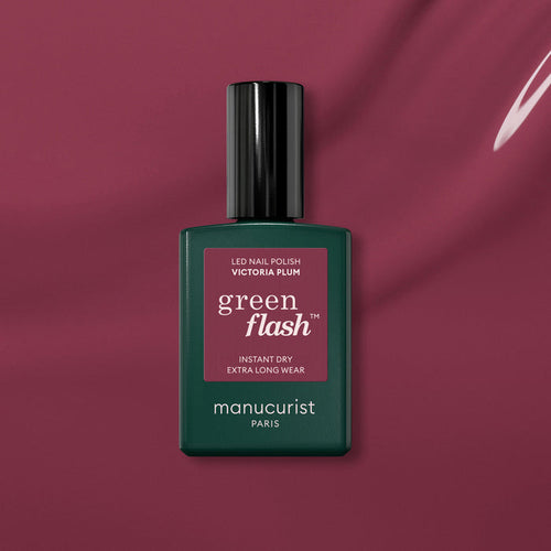 A bottle of Manucurist Victoria Plum gel polish sits on a matching plum background. The sleek dark green bottle has a maroon label and black cap, perfectly highlighting this rich plum shade.