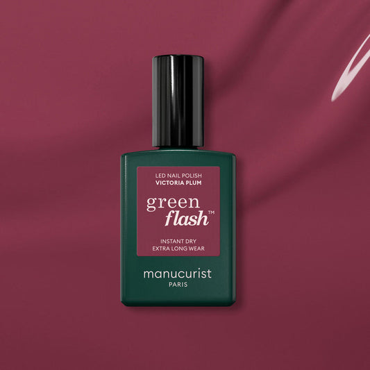 A bottle of Manucurist Victoria Plum gel polish sits on a matching plum background. The sleek dark green bottle has a maroon label and black cap, perfectly highlighting this rich plum shade.