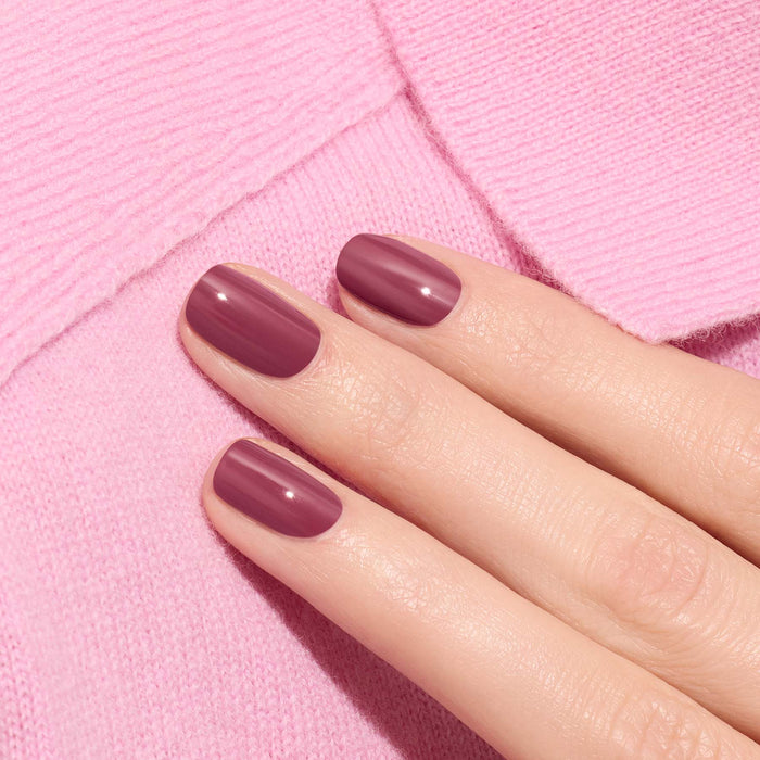 A hand with neatly manicured nails painted in glossy Manucurist Victoria Plum nail polish rests on soft pink textured fabric.