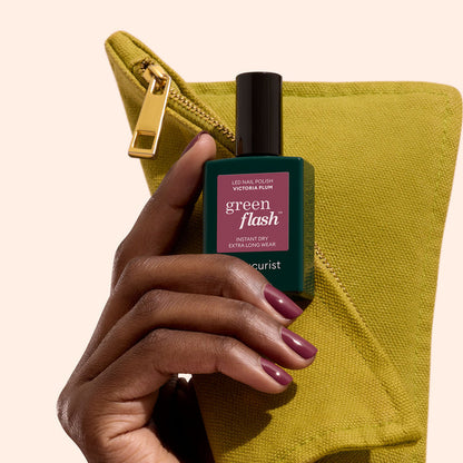 A hand with Manucurist Victoria Plum nails holds a bottle of gel polish in front of a yellow-green zippered pouch.